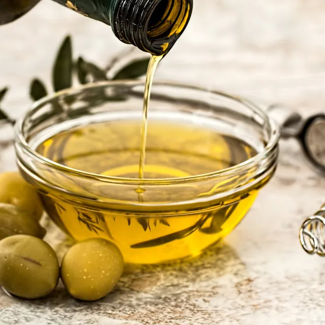 Extra virgin olive oil being poured from a bottle into a glass bowl, with green olives, an olive branch and kitchen tools alongside